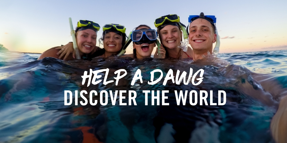 Five young people smiling in snorkeling gear, taking a selfie in blue water with a sunset in the background. Text: 'Help a dawg discover the world'